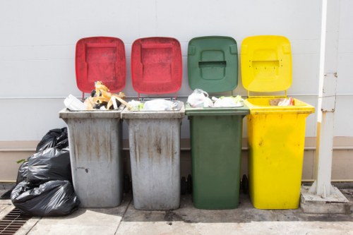 Four large wheelie bins made of hard plastic are positioned side by side on a paved surface in front of a white wall, with their lids open to reveal mixed rubbish inside. The first bin, in grey, contains various waste items, and next to it on the left are several tightly tied black plastic rubbish bags. The second bin, also grey, has visible waste spilling slightly from its open lid. The third bin is green, with its lid slightly ajar, and appears to contain mixed refuse as well. The fourth bin, in bright yellow, is positioned at the far right and is filled with rubbish, with some waste materials protruding from the open lid. The scene suggests a typical waste collection area in an urban or suburban setting, possibly associated with a commercial premises or residential street in Alperton, London, relevant to rubbish removal services offered by Office Clearance Alperton. The overall environment is clean and orderly, with minimal surrounding clutter, emphasizing the functional purpose of waste disposal bins only.