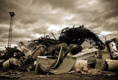 A large pile of discarded electronic appliances, metal debris, and twisted wires is situated outdoors on a flat, dirt ground, with some objects leaning and overlapping each other. The items include what appears to be old washing machines, metal cabinets, and various scrap metal parts, all showing signs of rust, dirt, and weathering. Behind the heap, a tangled mass of dark, thick cables or branches extends upward, contrasting with the lighter cloud-filled sky overhead, which has shades of grey and hints of sunlight breaking through. To the left in the background, a tall metal telecommunications tower is visible, indicating an urban or semi-urban area. The scene depicts a typical waste collection or clearance site, possibly in the vicinity of Alperton or nearby London, where Office Clearance Alperton might operate. The overall environment appears dusty and cluttered, emphasizing the nature of rubbish removal and clearance services for large-scale or industrial waste disposal.