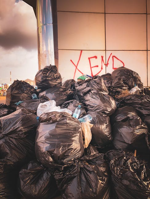 A person standing outdoors on a grassy area is holding open a large black rubbish bag with their gloved hands, preparing for waste disposal. The individual is dressed in a casual outfit, including a c
