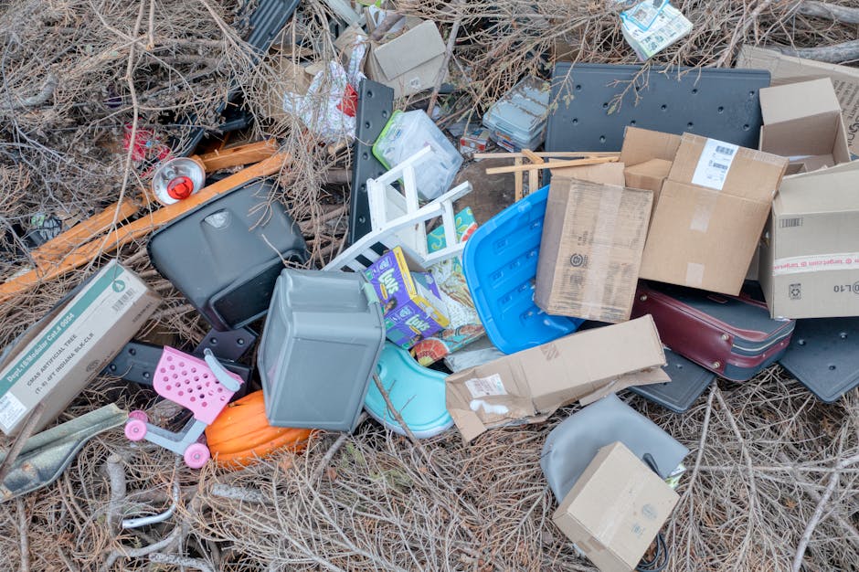 An aerial view of a large area covered with scattered waste and debris, including plastic bags, cardboard, and other miscellaneous refuse, situated next to a narrow footpath that runs parallel to a de