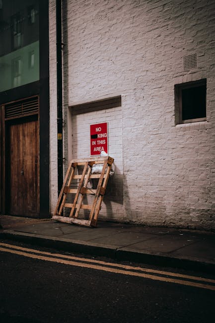 A person dressed in light-colored clothing is stacking a cardboard box on top of other similar boxes in an indoor setting, likely a room prepared for rubbish removal or a move. The cardboard boxes, va