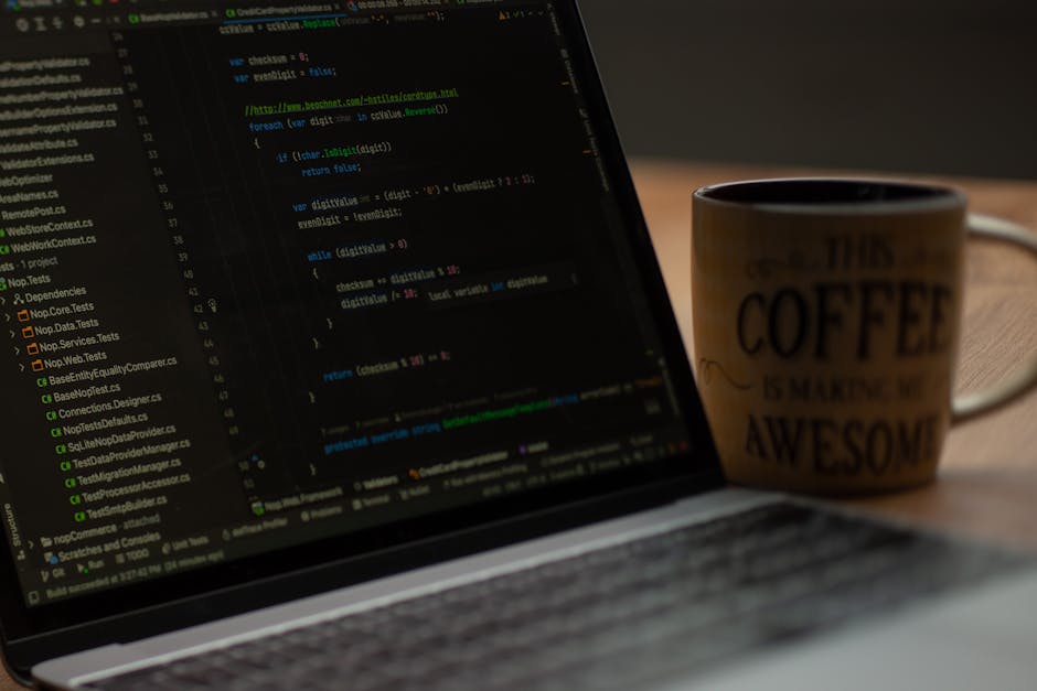 A close-up image of a laptop with a dark screen displaying lines of green and white code typical of programming or software development, positioned on a wooden surface. Next to the laptop, slightly be